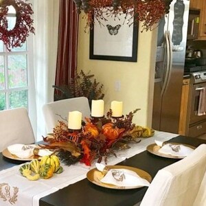 May include: A table setting for a Thanksgiving dinner with a white tablecloth, gold chargers, and white napkins. The centerpiece features a variety of fall foliage, gourds, and candles.