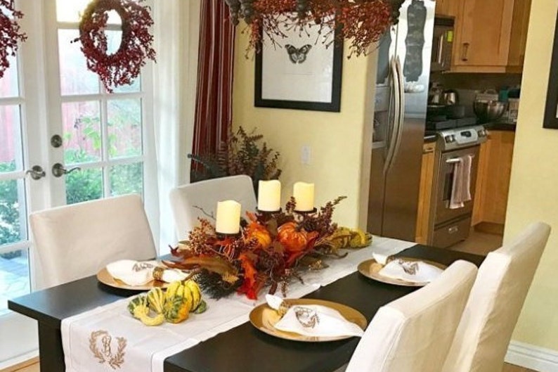 May include: A table setting for a Thanksgiving dinner with a white tablecloth, gold chargers, and white napkins. The centerpiece features a variety of fall foliage, gourds, and candles.