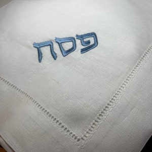 Może przedstawiać: Biała serwetka lniana z wyszytym niebieskim hebrajskim słowem "Pesach". Serwetka ma obszyty brzeg.