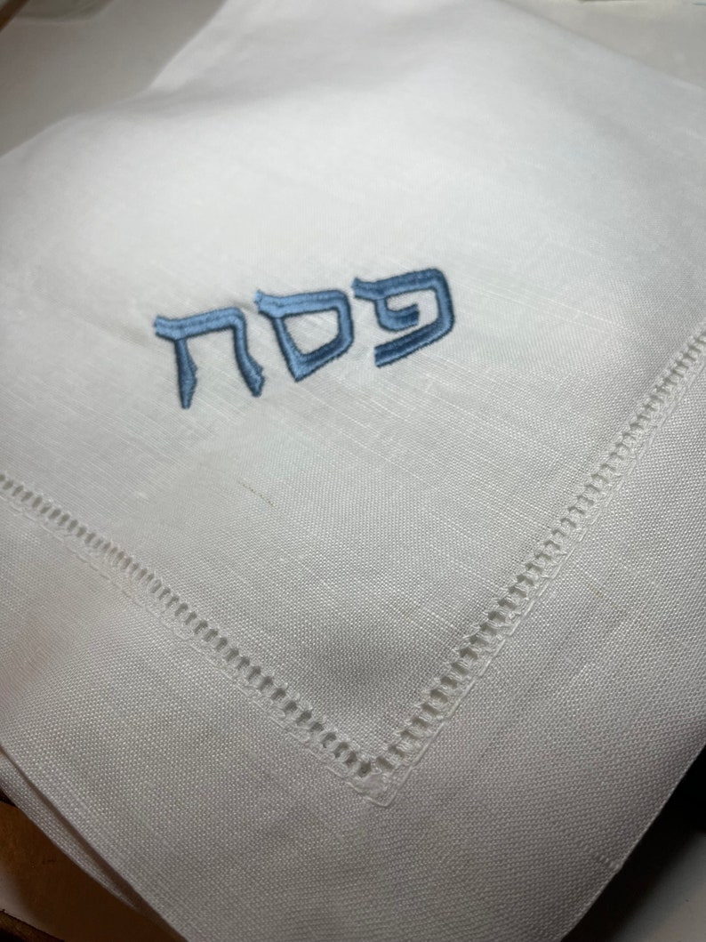 Może przedstawiać: Biała serwetka lniana z wyszytym niebieskim hebrajskim słowem "Pesach". Serwetka ma obszyty brzeg.