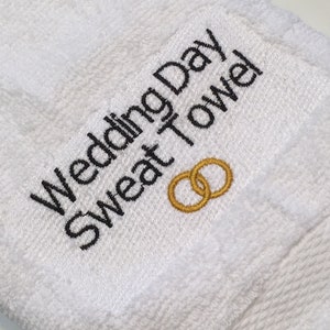 K&ouml;nnte beinhalten: Wei&szlig;es Frottee-Schwei&szlig;handtuch mit schwarzem gesticktem Text "Wedding Day Sweat Towel". Zwei goldene Eheringe sind darunter gestickt. Ein Accessoire zum Thema Hochzeit.