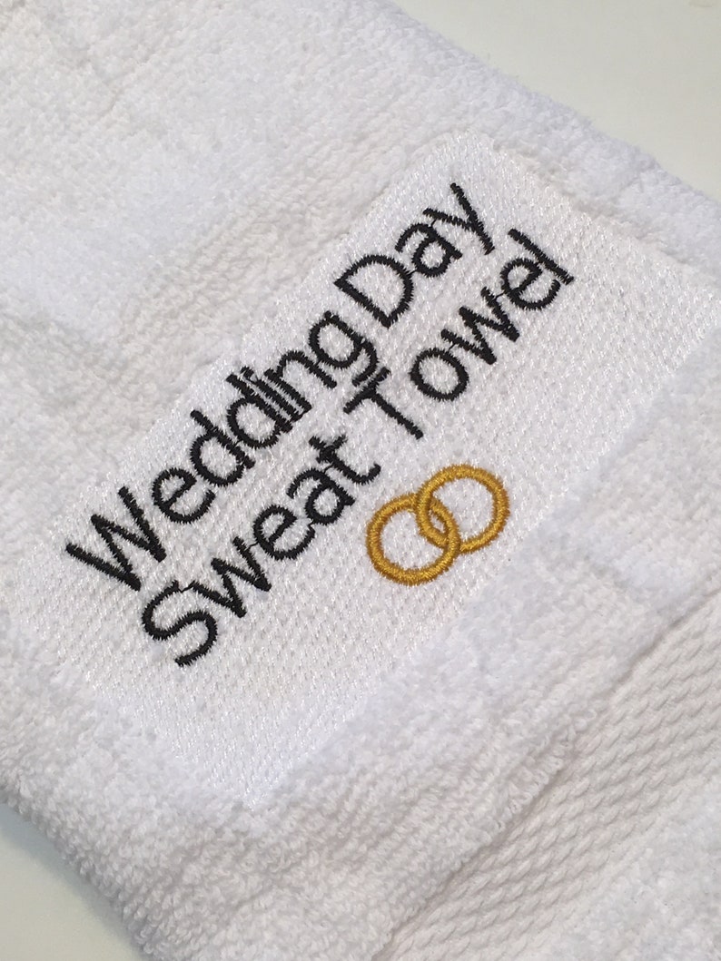 K&ouml;nnte beinhalten: Wei&szlig;es Frottee-Schwei&szlig;handtuch mit schwarzem gesticktem Text "Wedding Day Sweat Towel". Zwei goldene Eheringe sind darunter gestickt. Ein Accessoire zum Thema Hochzeit.
