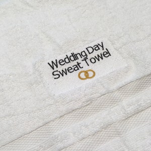 K&ouml;nnte beinhalten: Wei&szlig;es Frotteehandtuch mit dem gestickten Text "Wedding Day Sweat Towel" in Schwarz. Unter dem Text befinden sich zwei ineinandergreifende goldene Ringe. Das Handtuch ist ein helles, leuchtendes Wei&szlig;.