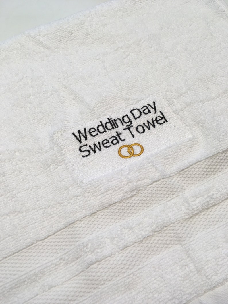 K&ouml;nnte beinhalten: Wei&szlig;es Frotteehandtuch mit dem gestickten Text "Wedding Day Sweat Towel" in Schwarz. Unter dem Text befinden sich zwei ineinandergreifende goldene Ringe. Das Handtuch ist ein helles, leuchtendes Wei&szlig;.