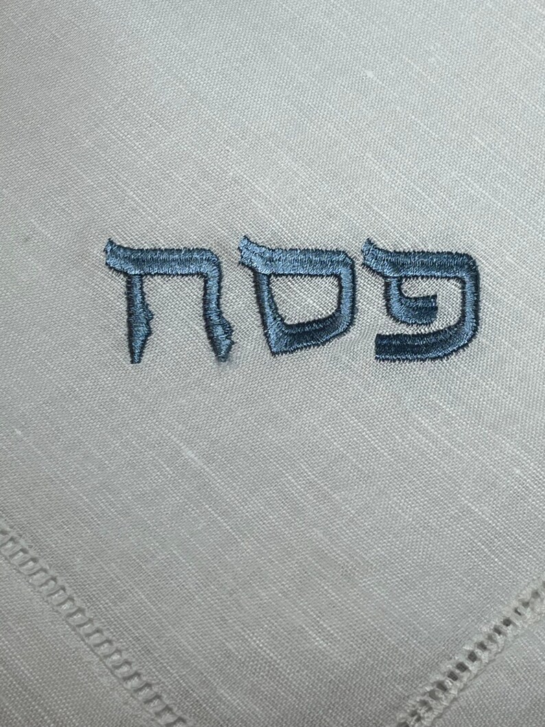 Może przedstawiać: Biała serwetka lniana z haftowanym niebieską nicią hebrajskim słowem "Pesach".