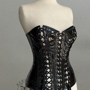 May include: Black patent leather corset with lace-up detailing and a sweetheart neckline. The corset is displayed on a mannequin and features a logo that says "ELEGANCE FASHION ART".
