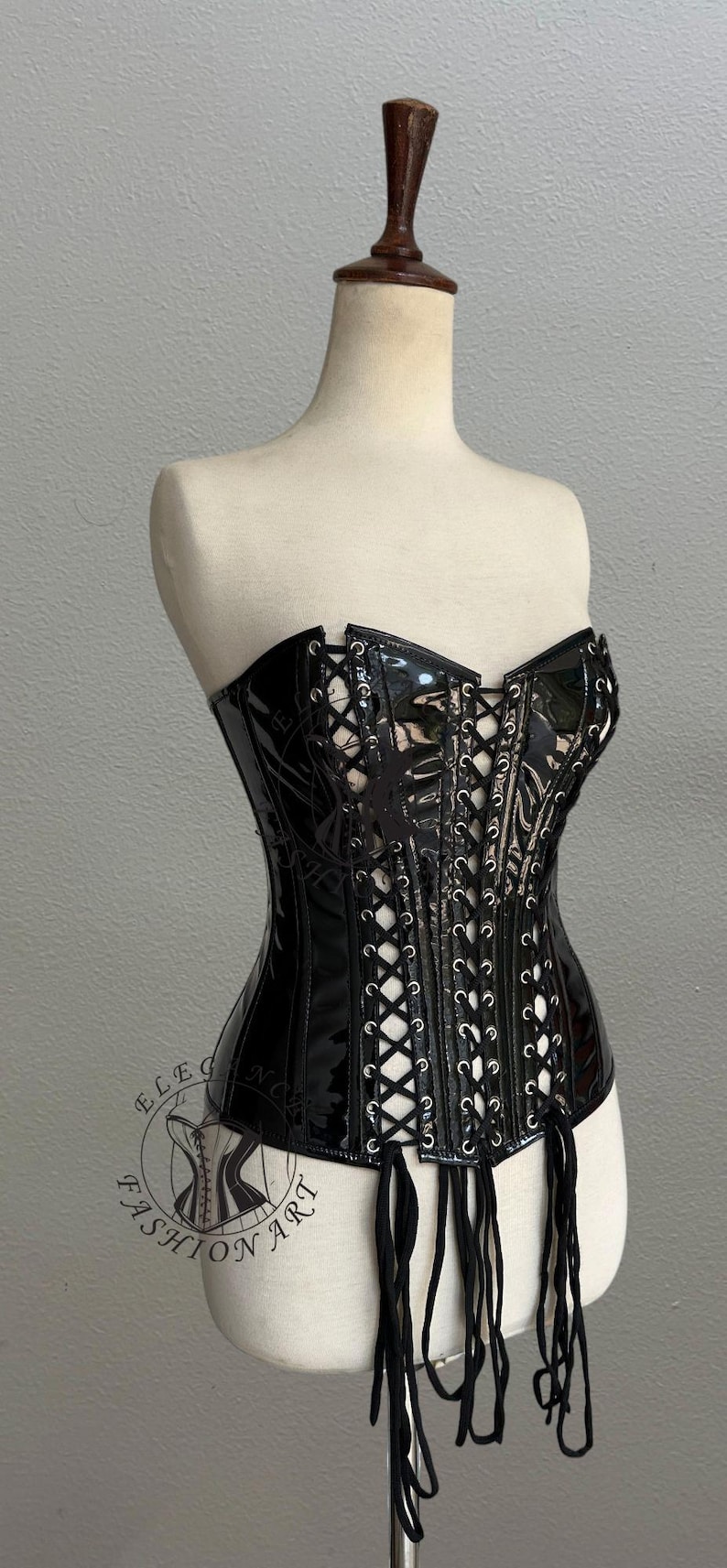 May include: Black patent leather corset with lace-up detailing and a sweetheart neckline. The corset is displayed on a mannequin and features a logo that says "ELEGANCE FASHION ART".