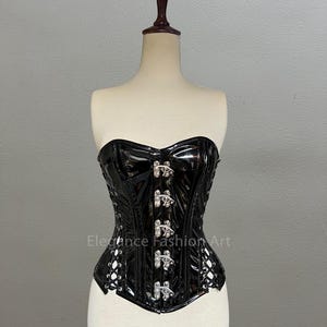 Pu&ograve; includere: Corsetto in pelle verniciata nera con fibbie argentate e schiena con lacci. Il corsetto &egrave; su un manichino bianco. Il testo "Elegance Fashion Art" &egrave; stampato sul davanti del corsetto.