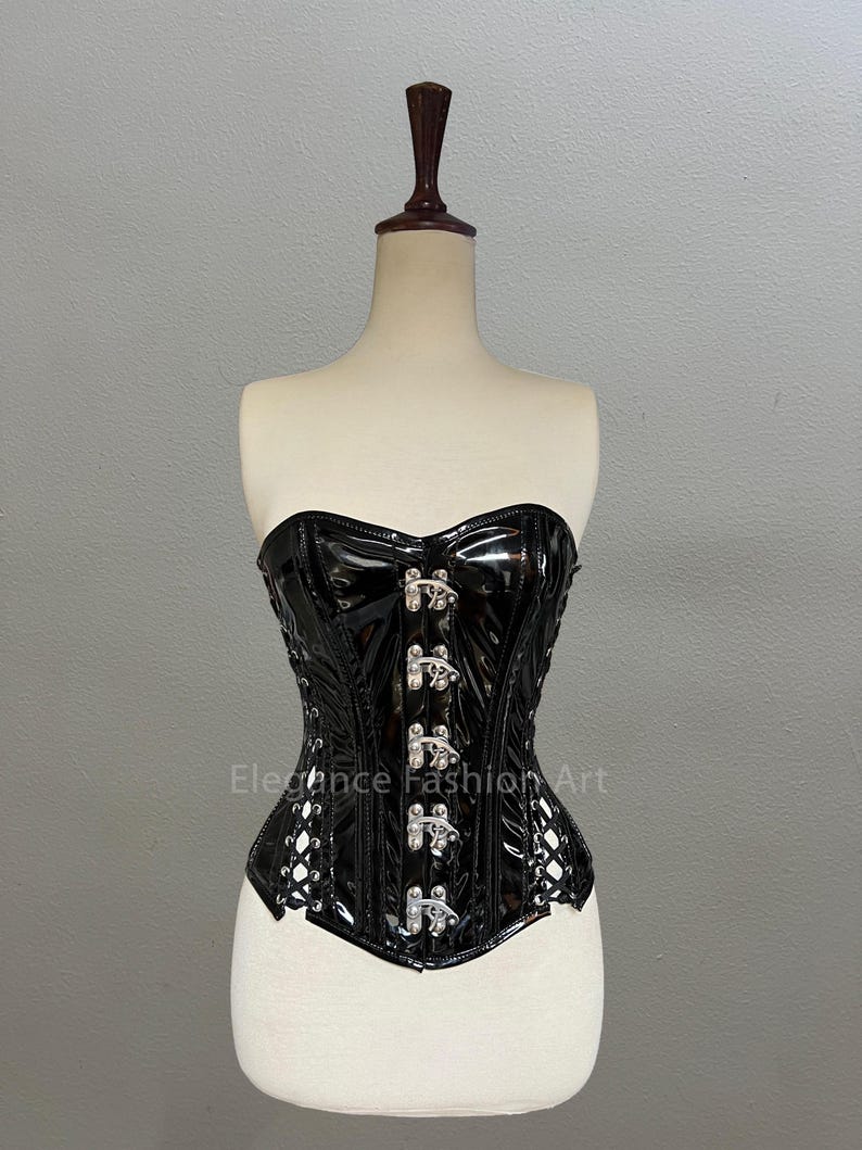 Pu&ograve; includere: Corsetto in pelle verniciata nera con fibbie argentate e schiena con lacci. Il corsetto &egrave; su un manichino bianco. Il testo "Elegance Fashion Art" &egrave; stampato sul davanti del corsetto.