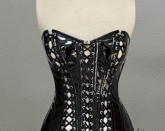 Op de afbeelding: Zwart, strapless korset met een glanzende afwerking, met een veter aan de voorkant en decoratieve uitsparingen. Het korset heeft bevestigde zwarte bandjes en wordt op een mannequin getoond. Modeartikel.
