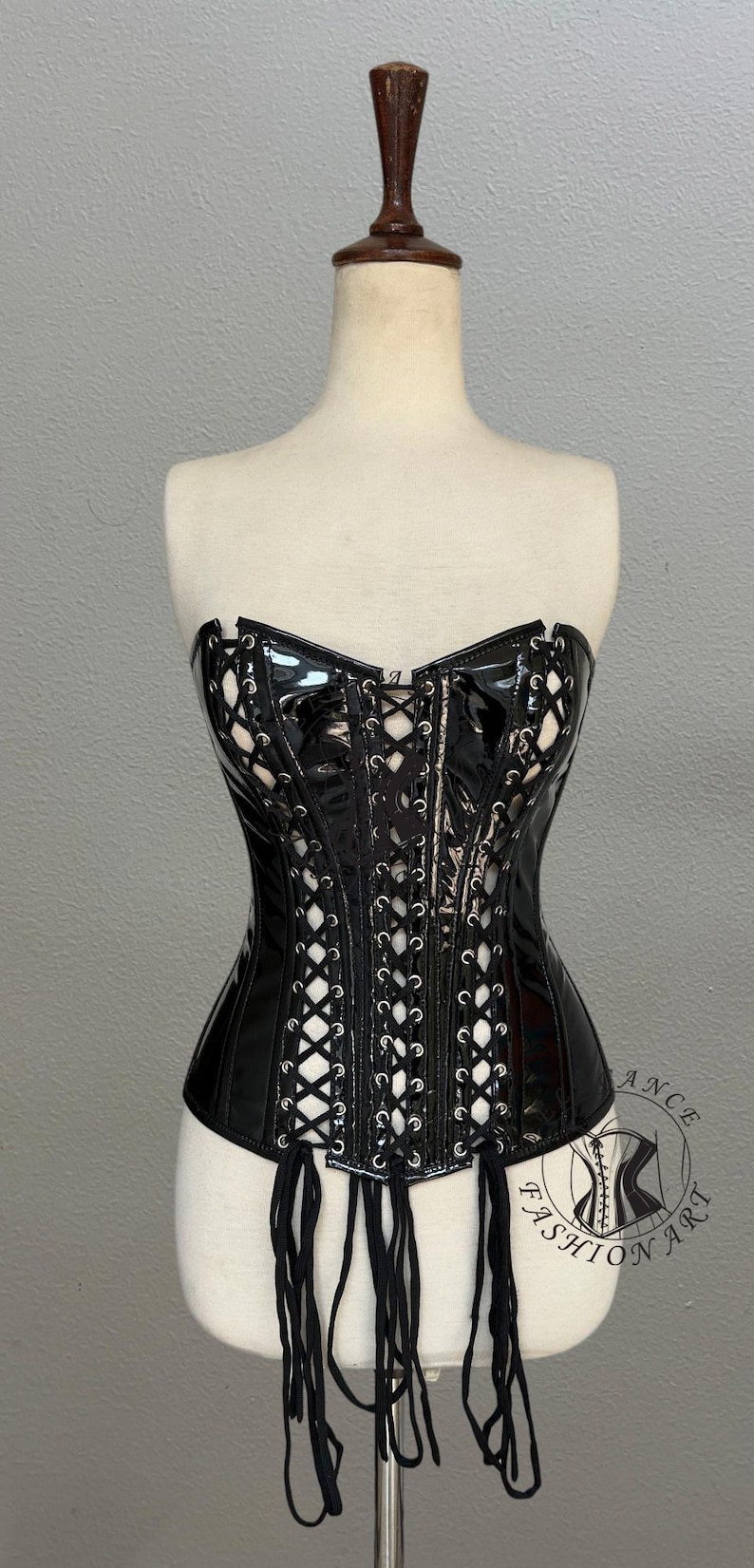 May include: Black, strapless corset with a glossy finish, featuring a lace-up front and decorative cutouts. The corset has attached black straps and is displayed on a mannequin. The corset is a fashion item.