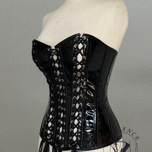May include: A black, glossy corset with white lacing and decorative fringe. The corset is displayed on a mannequin. The garment has a structured silhouette and a high-shine finish, typical of a fashion item.
