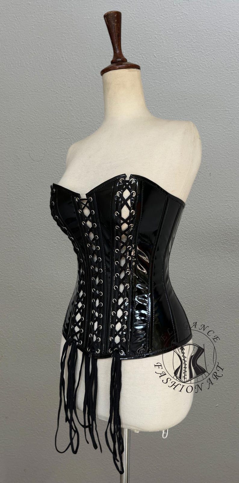 May include: A black, glossy corset with white lacing and decorative fringe. The corset is displayed on a mannequin. The garment has a structured silhouette and a high-shine finish, typical of a fashion item.