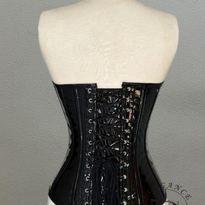 May include: A black, glossy corset with a lace-up back, displayed on a mannequin. The corset has a structured design with visible boning and a slight curve at the waist. The back features a series of eyelets for lacing.