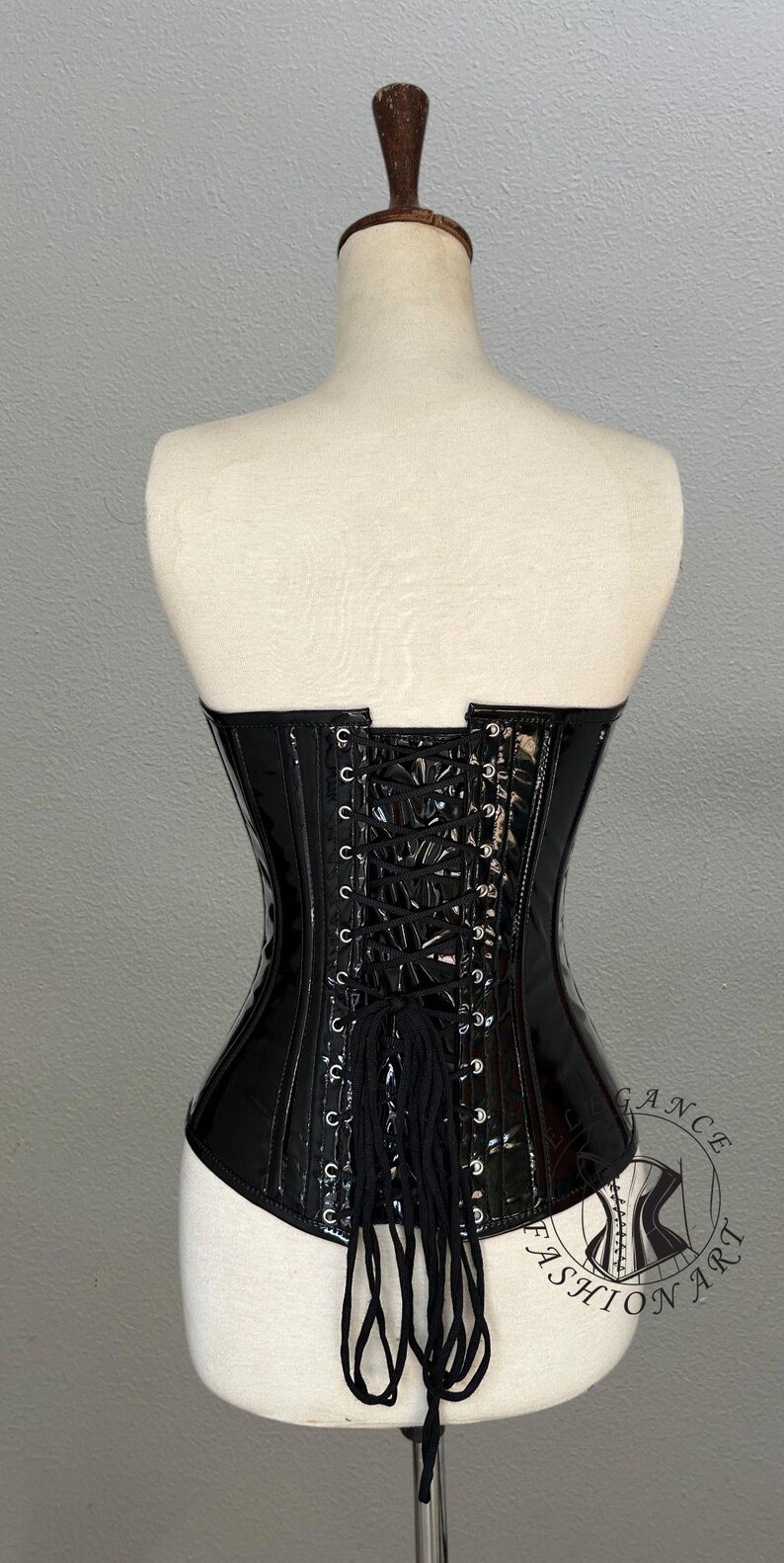 May include: A black, glossy corset with a lace-up back, displayed on a mannequin. The corset has a structured design with visible boning and a slight curve at the waist. The back features a series of eyelets for lacing.