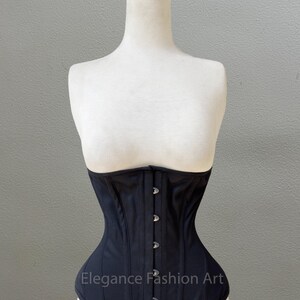 May include: A black corset with a satin finish and silver metal clasps. The corset is on a white mannequin. The text "Elegance Fashion Art" is printed on the front of the corset.
