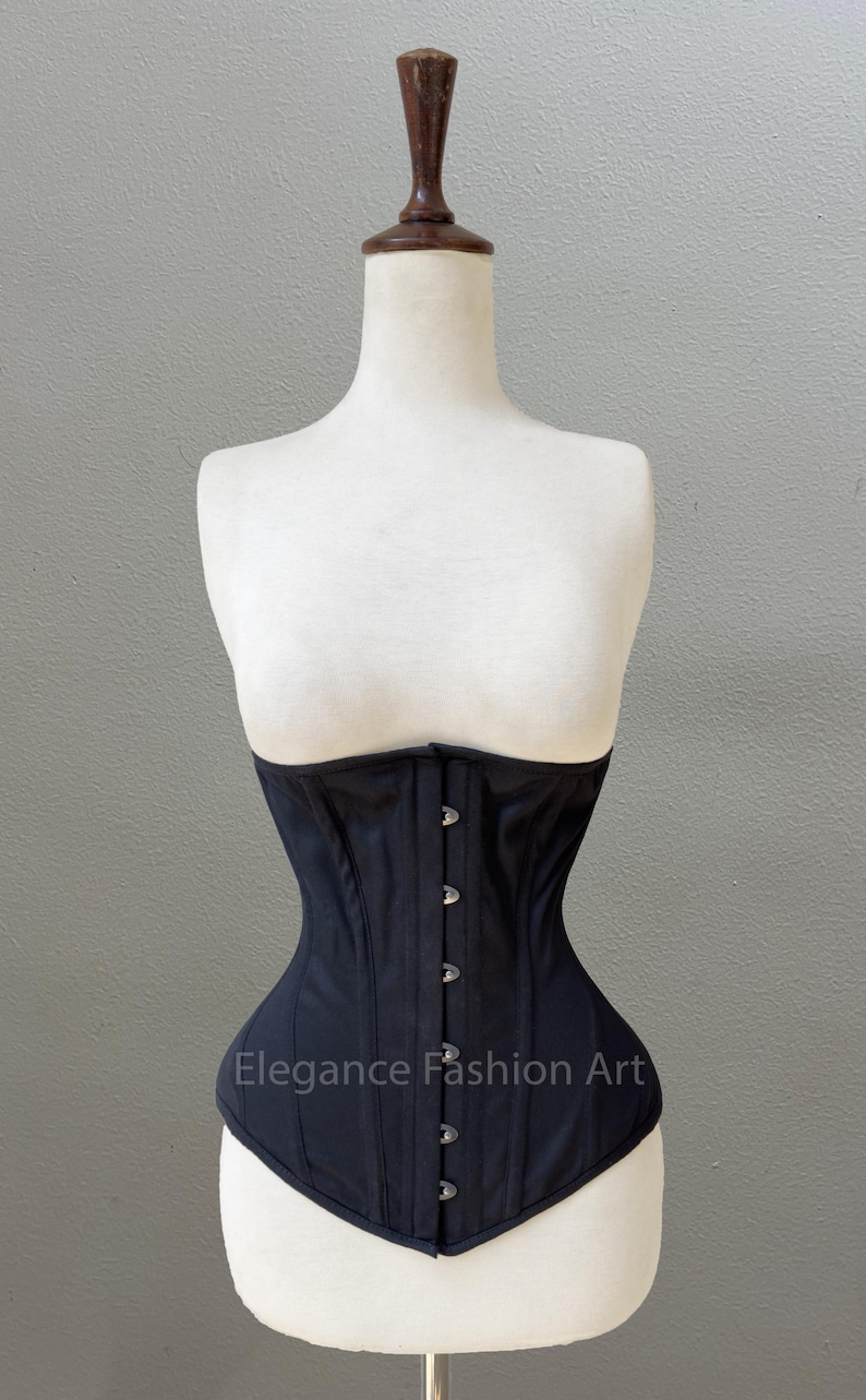 May include: A black corset with a satin finish and silver metal clasps. The corset is on a white mannequin. The text "Elegance Fashion Art" is printed on the front of the corset.