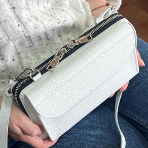 May include: A white rectangular crossbody bag with a pebbled leather texture, featuring a front flap closure and a black zippered compartment along the top. The bag includes a matching white adjustable shoulder strap and silver-toned metal hardware.