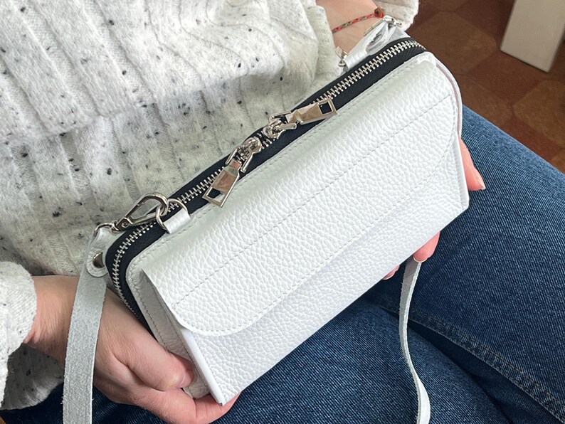 May include: A white rectangular crossbody bag with a pebbled leather texture, featuring a front flap closure and a black zippered compartment along the top. The bag includes a matching white adjustable shoulder strap and silver-toned metal hardware.