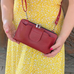 Könnte beinhalten: Eine rote Umhängetasche aus genarbtem Leder in rechteckiger Form mit einer Fronttasche mit Klappe und geschwungenem Rand sowie einem Reißverschluss oben. Die Tasche hat einen passenden, verstellbaren Schulterriemen mit silberfarbenen Metallbeschlägen.