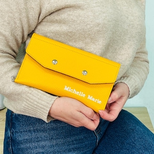 May include: A bright yellow clutch with a textured surface, secured by two silver snap closures. The name "Michelle Marie" is embroidered in white on the front. The clutch is held against a background of blue jeans and a beige sweater.