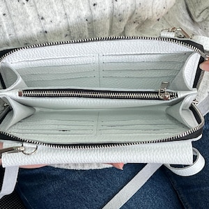 May include: A white textured crossbody bag with a zippered main compartment, multiple interior card slots, and a central zippered pocket. The bag features silver-toned metal hardware, a matching white adjustable strap, and a structured rectangular shape.