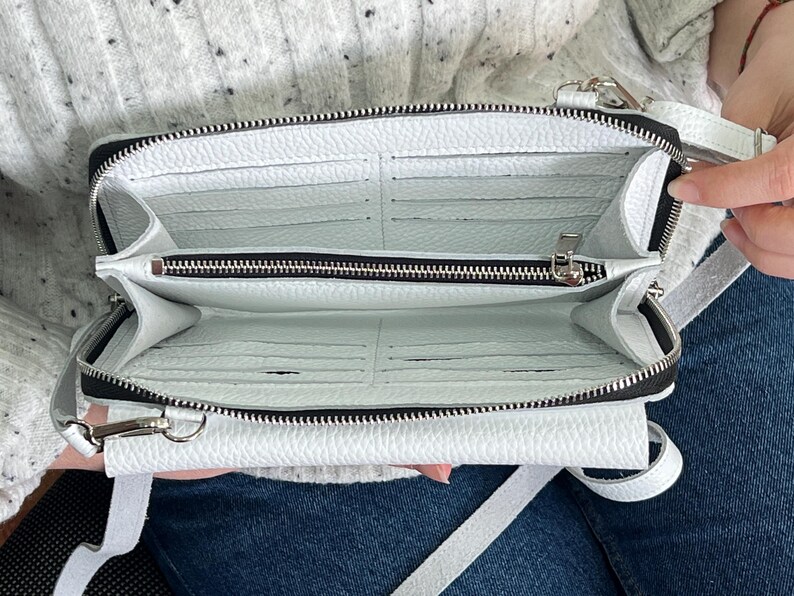 May include: A white textured crossbody bag with a zippered main compartment, multiple interior card slots, and a central zippered pocket. The bag features silver-toned metal hardware, a matching white adjustable strap, and a structured rectangular shape.