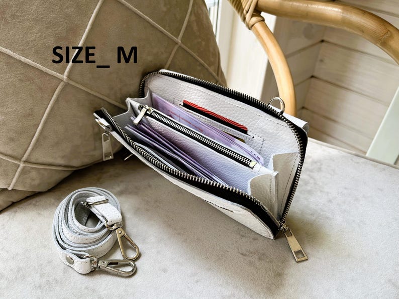 May include: A white textured leather crossbody bag with a black zipper closure and silver-toned hardware. The interior features multiple compartments containing paper currency and cards. The text SIZE_M is visible. A detachable white leather strap is included.