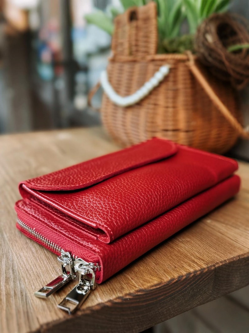 May include: A vibrant red wallet with a textured, pebbled surface. The wallet features a flap closure on the top section and a separate compartment secured by two silver-toned metal zippers with rectangular pulls, positioned on the side.