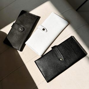 May include: Three rectangular wallets with a textured surface and a snap button closure. Two of the wallets are black and one is white. Each wallet features a flap that secures with a silver-toned metal snap, providing a clean and functional design.