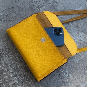 May include: A bright yellow textured leather crossbody bag with a flap closure secured by a silver-toned magnetic snap. The bag features a matching yellow shoulder strap and contains a dark blue smartphone tucked inside the open front pocket.