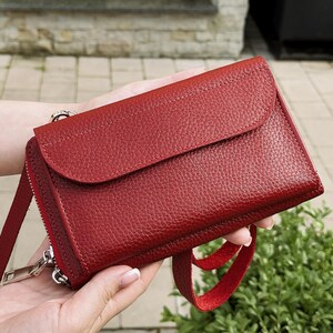 May include: A deep red leather crossbody bag with a textured pebble grain finish. The rectangular bag features a front flap closure, a side zipper with a silver-toned metal pull, and a matching red leather strap attached to the side hardware.