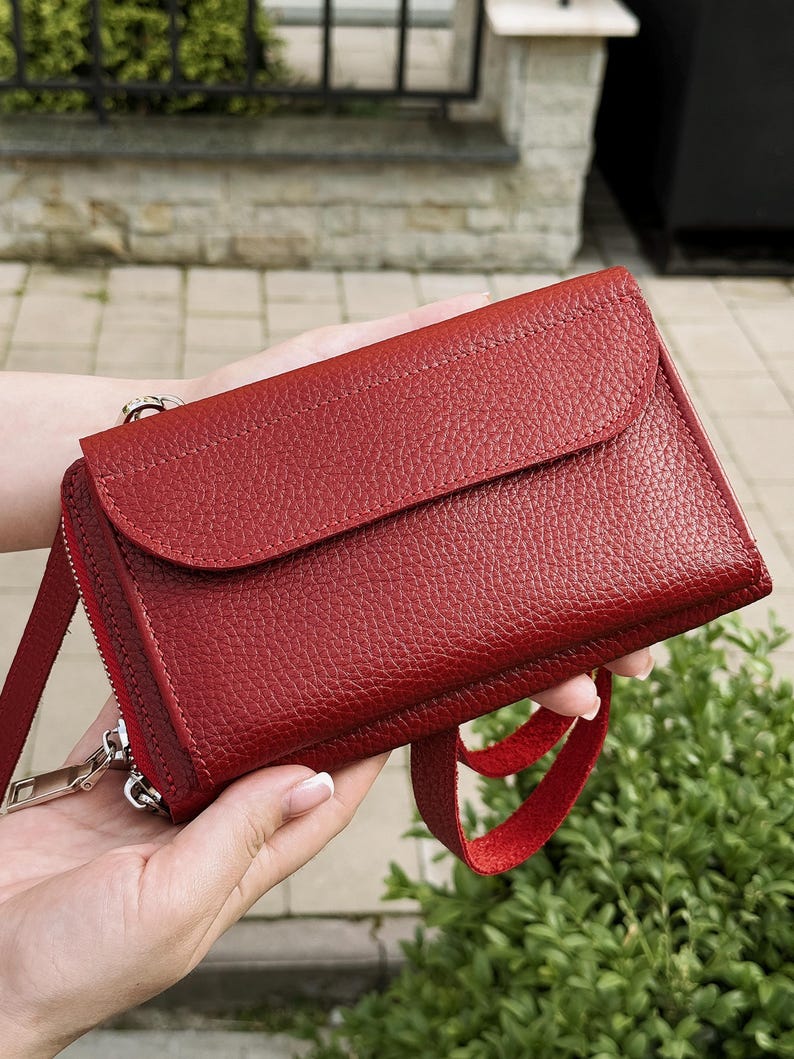 May include: A deep red leather crossbody bag with a textured pebble grain finish. The rectangular bag features a front flap closure, a side zipper with a silver-toned metal pull, and a matching red leather strap attached to the side hardware.