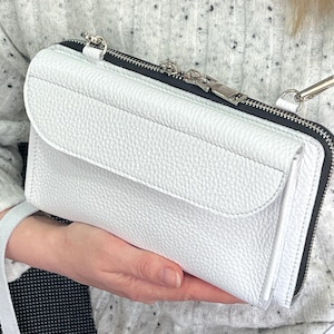 May include: A white pebbled leather crossbody bag featuring a rectangular silhouette, a front flap pocket, and a top zipper closure with silver-toned metal hardware. The bag includes a matching white leather shoulder strap attached with silver clips.