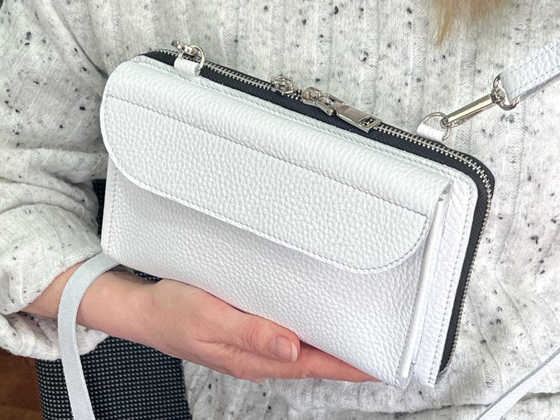 May include: A white pebbled leather crossbody bag featuring a rectangular silhouette, a front flap pocket, and a top zipper closure with silver-toned metal hardware. The bag includes a matching white leather shoulder strap attached with silver clips.