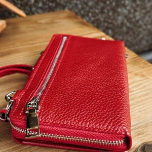 May include: A rectangular red leather wallet with a pebbled texture, featuring a silver-toned metal zipper closure along the top edge and a matching silver-toned zipper pull on a side pocket. The wallet has a structured design with visible stitching.