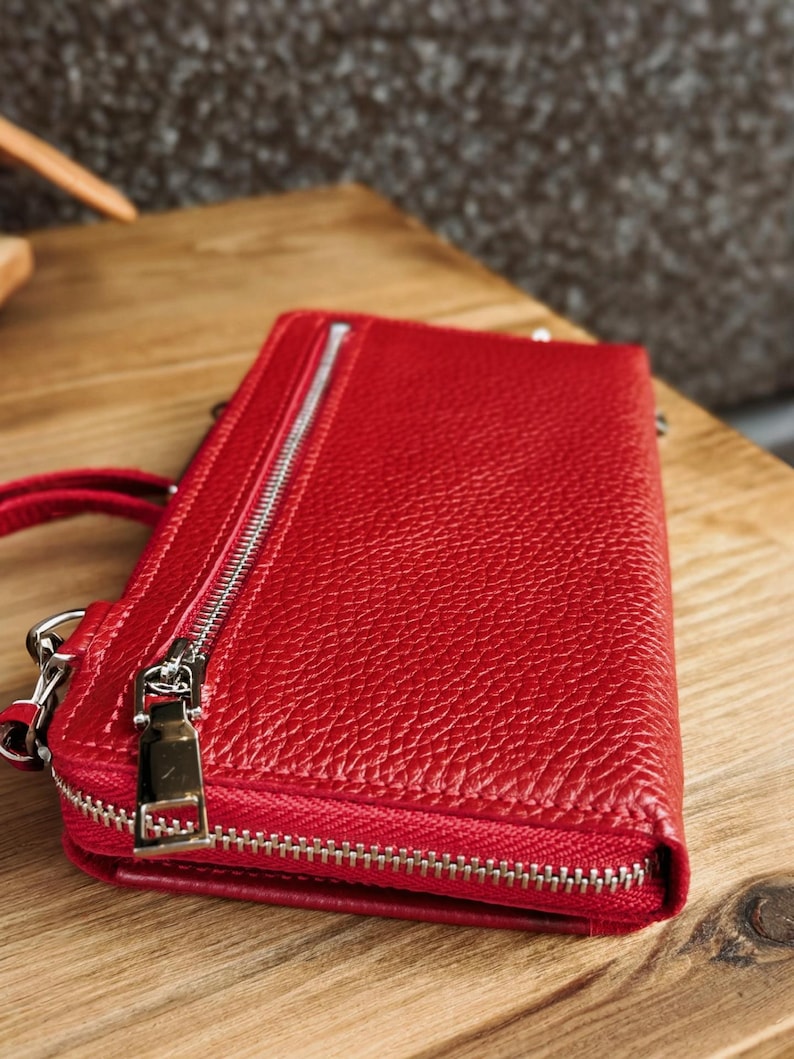 May include: A rectangular red leather wallet with a pebbled texture, featuring a silver-toned metal zipper closure along the top edge and a matching silver-toned zipper pull on a side pocket. The wallet has a structured design with visible stitching.