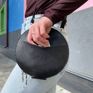 Op de afbeelding: Een zwarte ronde schoudertas met een ritssluiting en een zilveren kettingdetail. De tas is gemaakt van gestructureerd leer en heeft een klep met een zak.