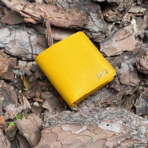 May include: A bright yellow leather wallet with a textured surface. The wallet has the initials "MG" embossed on the front. The wallet is resting on a bed of brown and gray wood chips.