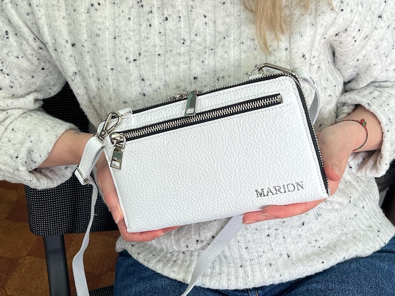 May include: A white rectangular crossbody bag with a pebbled texture, featuring a silver-toned zipper closure, a front zippered pocket, and a detachable shoulder strap. The name MARION is embossed in silver lettering on the lower right corner.