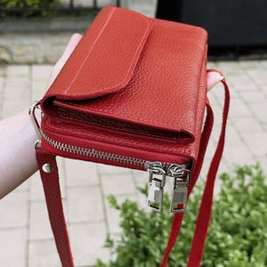 May include: A vibrant red leather crossbody bag with a textured pebble grain finish. The bag features a flap closure on the top section and a separate zippered compartment below with two silver-toned metal zipper pulls and a matching red leather strap.