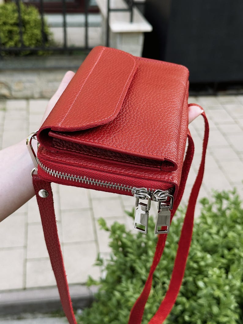 May include: A vibrant red leather crossbody bag with a textured pebble grain finish. The bag features a flap closure on the top section and a separate zippered compartment below with two silver-toned metal zipper pulls and a matching red leather strap.