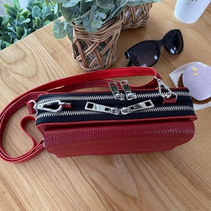 以下が含まれることがあります： 黒いジッパーパネルとシルバーの金具が付いた赤い革のクロスボディバッグ。バッグには長い赤いストラップとテクスチャード加工が施されています。サングラスとペストリーのある木製の表面にあります。