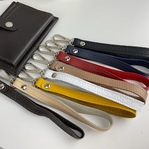 May include: A set of eight leather keychains in various colors, including black, navy blue, red, beige, white, and yellow. The keychains are made of leather and have a silver metal clasp.