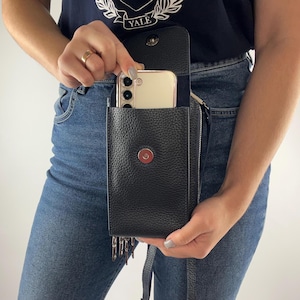 Może przedstawiać: Czarna skórzana torba na telefon na ramię z magnetycznym zapięciem. Torba ma teksturowane wykończenie, długi pasek i mały uchwyt na klucze. Smartfon jest widoczny w otwartej komorze.