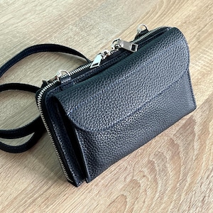 May include: A black crossbody bag with a pebbled texture, featuring a front flap closure, a top zipper, and a thin matching shoulder strap. The bag has silver-toned metal hardware, including zipper pulls and attachment rings for the strap.