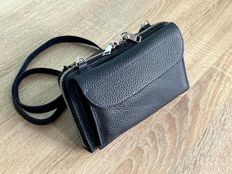 May include: A black crossbody bag with a pebbled texture, featuring a front flap closure, a top zipper, and a thin matching shoulder strap. The bag has silver-toned metal hardware, including zipper pulls and attachment rings for the strap.