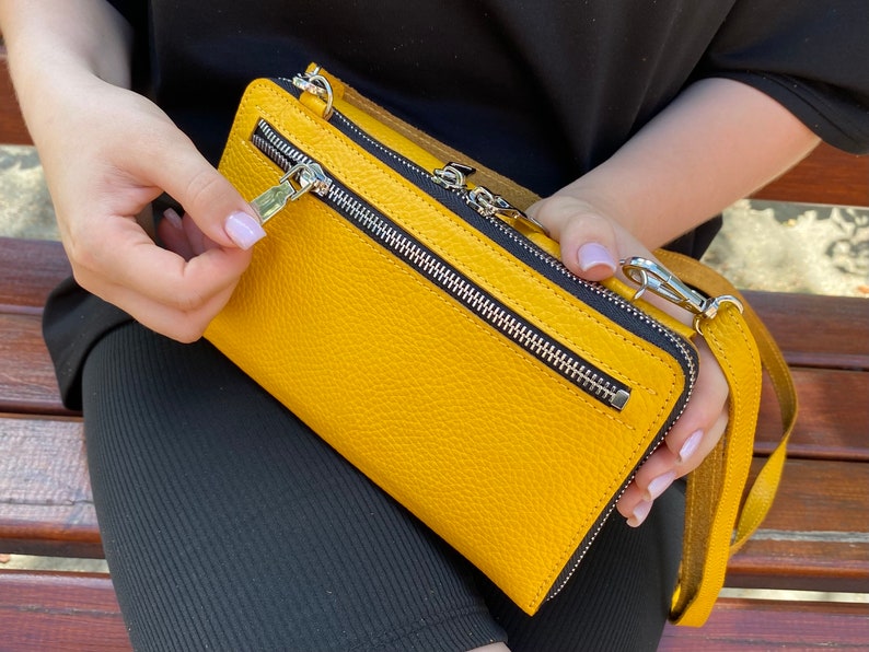 May include: A mustard yellow rectangular crossbody bag with a pebbled texture, featuring a prominent horizontal silver metal zipper on the front, a top zipper closure, and a matching yellow leather shoulder strap with silver metal swivel clips.