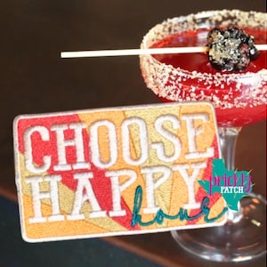 May include: A colorful embroidered patch with the words "CHOOSE HAPPY hour" in white and teal stitching. The patch is rectangular with a red, yellow, and orange background. A cocktail with a sugared rim and a blackberry garnish is in the background.