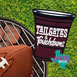 May include: A maroon embroidered patch shaped like a cup with a black rim, featuring the white text "TAILGATES & TOUCHDOWNS". The patch is positioned near a brown textured football and a metal grill grate, with a teal logo reading "prickly PATCH".